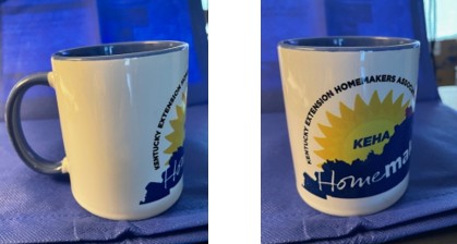 A white ceramic mug with blue insides and handle. On the mug is the Kentucky Extension Homemaker Association Logo, with the letters KEHA inside a sun rising over an image of the state with the word Homemakers inside.